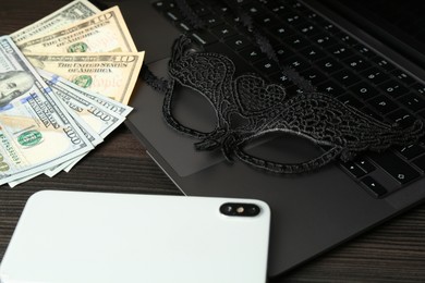 Photo of Sex work. Laptop, mask, smartphone and money on wooden surface, closeup