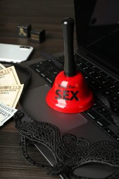 Sex work. Laptop, mask, money, smartphone, dice and bell on wooden surface, closeup