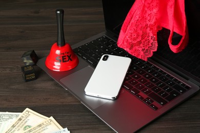 Photo of Sex work. Laptop with lingerie, smartphone, bell, dice and money on wooden surface, closeup