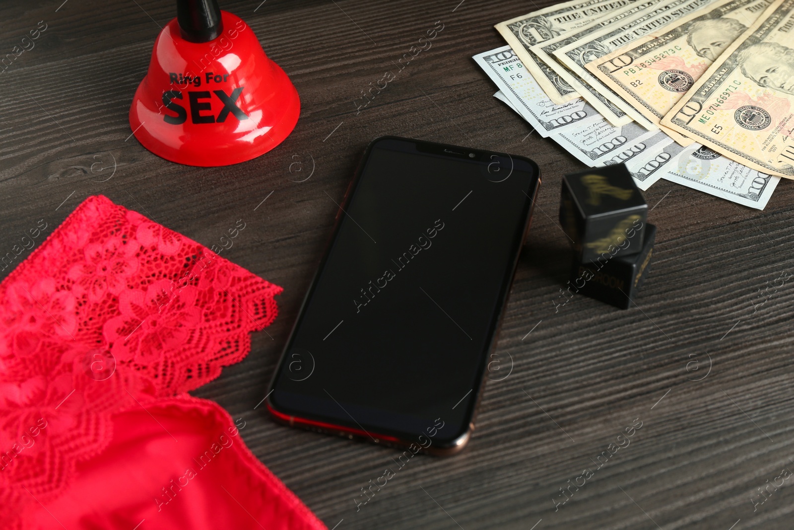 Photo of Sex work. Smartphone, underwear, bell, dice and money on wooden surface, closeup