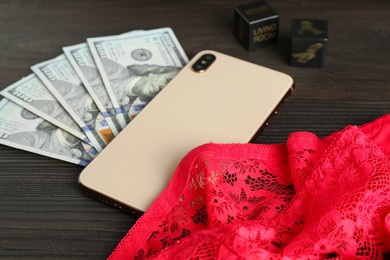 Photo of Sex work. Smartphone, underwear, dice and money on wooden surface, closeup