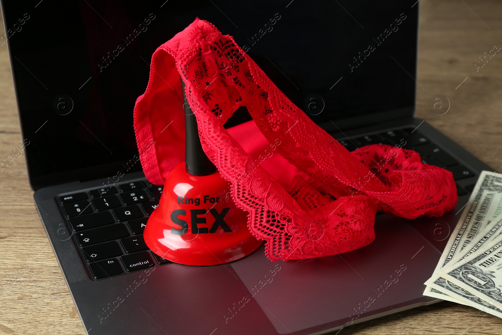 Photo of Sex work. Laptop with lingerie, bell and money on wooden surface, closeup