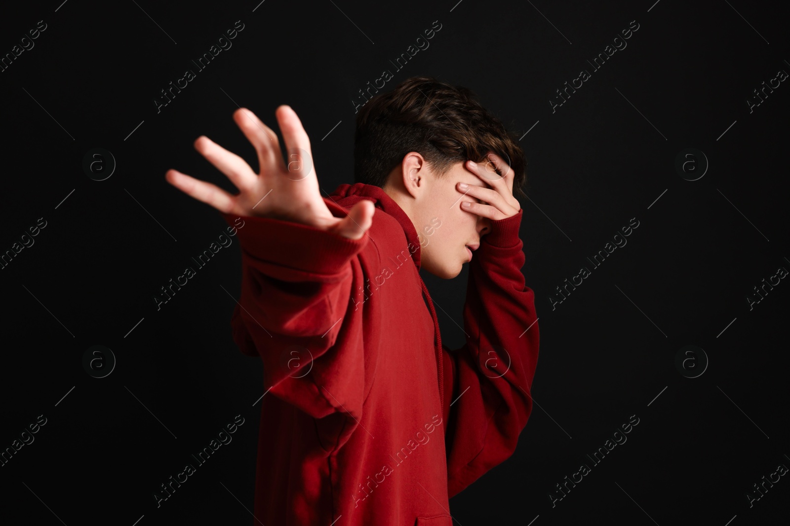 Photo of Scared teenage boy covering face with hands on black background