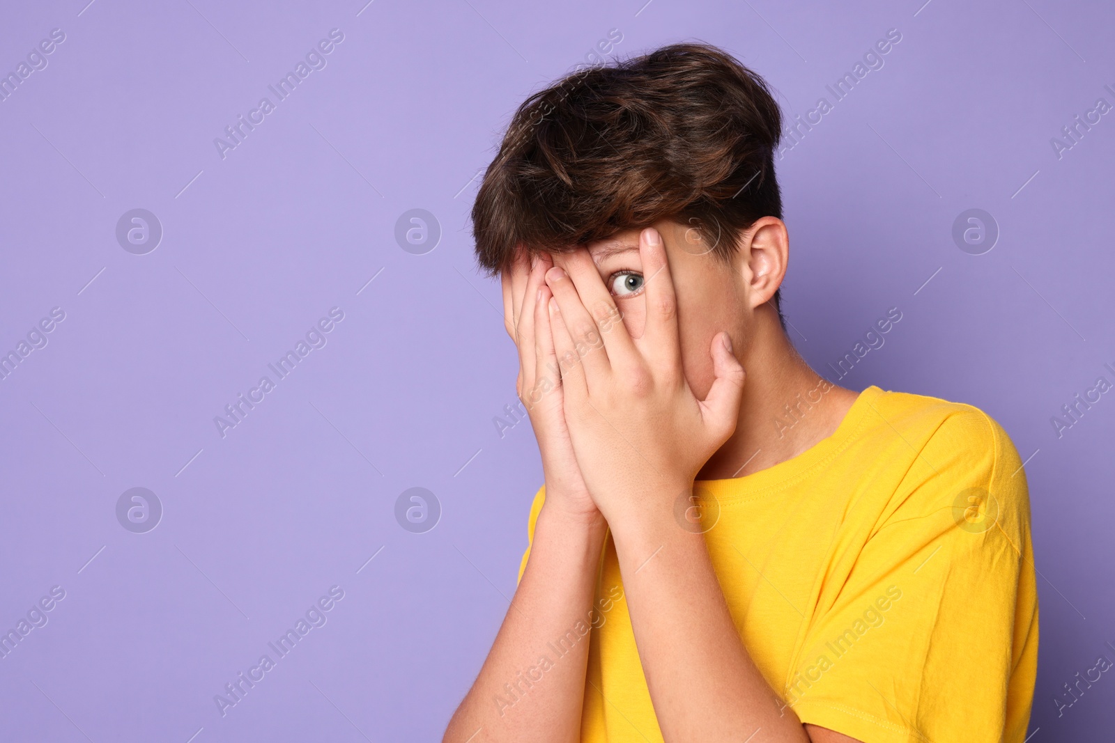 Photo of Scared teenage boy covering face with hands on violet background. Space for text