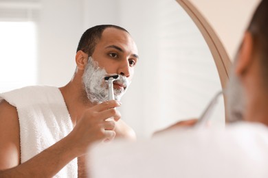 Photo of Handsome man shaving with razor near mirror in bathroom