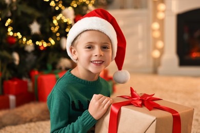 Photo of Cute little boy in Santa hat with Christmas gift indoors