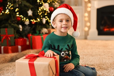 Photo of Cute little boy in Santa hat with Christmas gift indoors