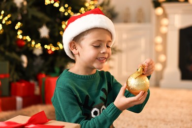 Photo of Cute little boy in Santa hat with Christmas ball at home