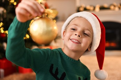 Photo of Cute little boy in Santa hat with Christmas ball at home