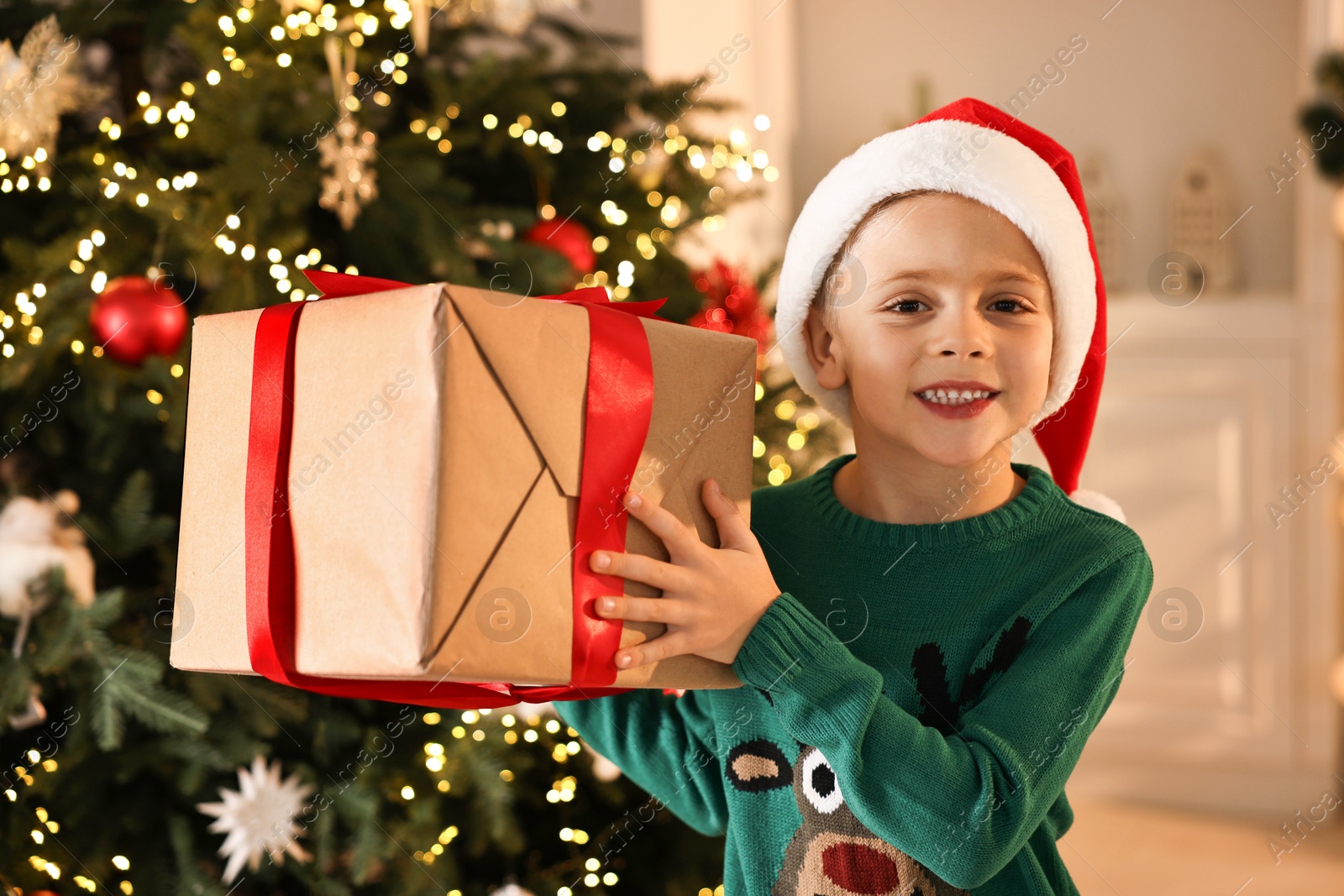 Photo of Cute little boy in Santa hat with Christmas gift indoors