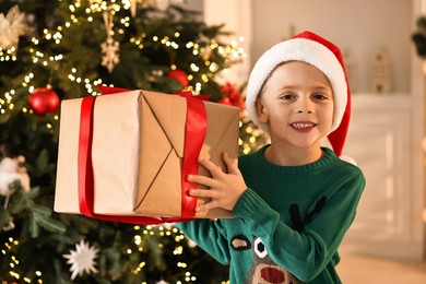 Photo of Cute little boy in Santa hat with Christmas gift indoors