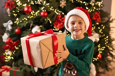 Photo of Cute little boy in Santa hat with Christmas gift indoors