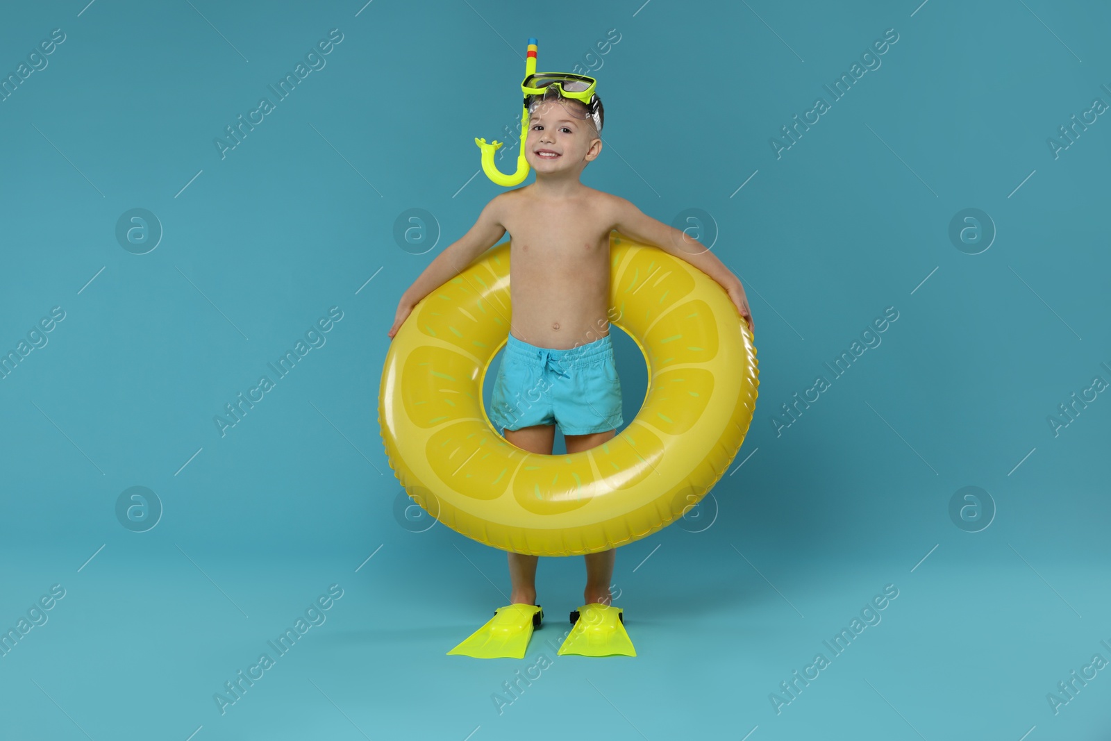 Photo of Cute little boy in beachwear with snorkeling equipment and inflatable ring on light blue background