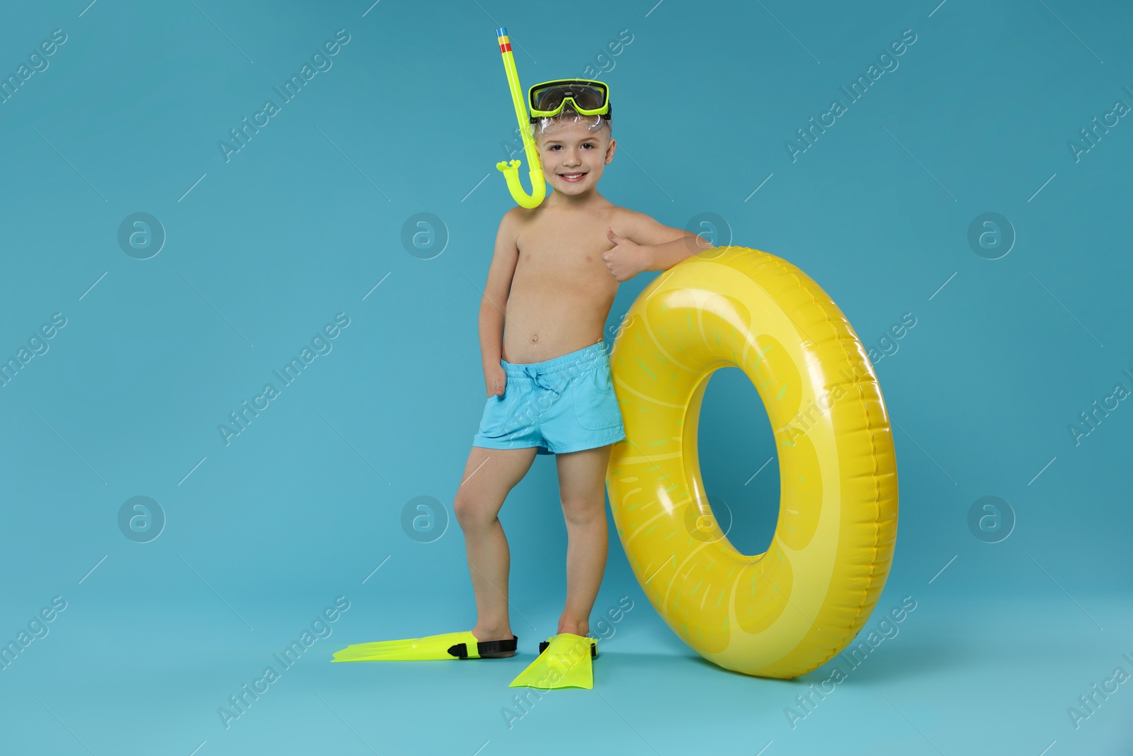 Photo of Cute little boy in beachwear with snorkeling equipment and inflatable ring showing thumbs up on light blue background