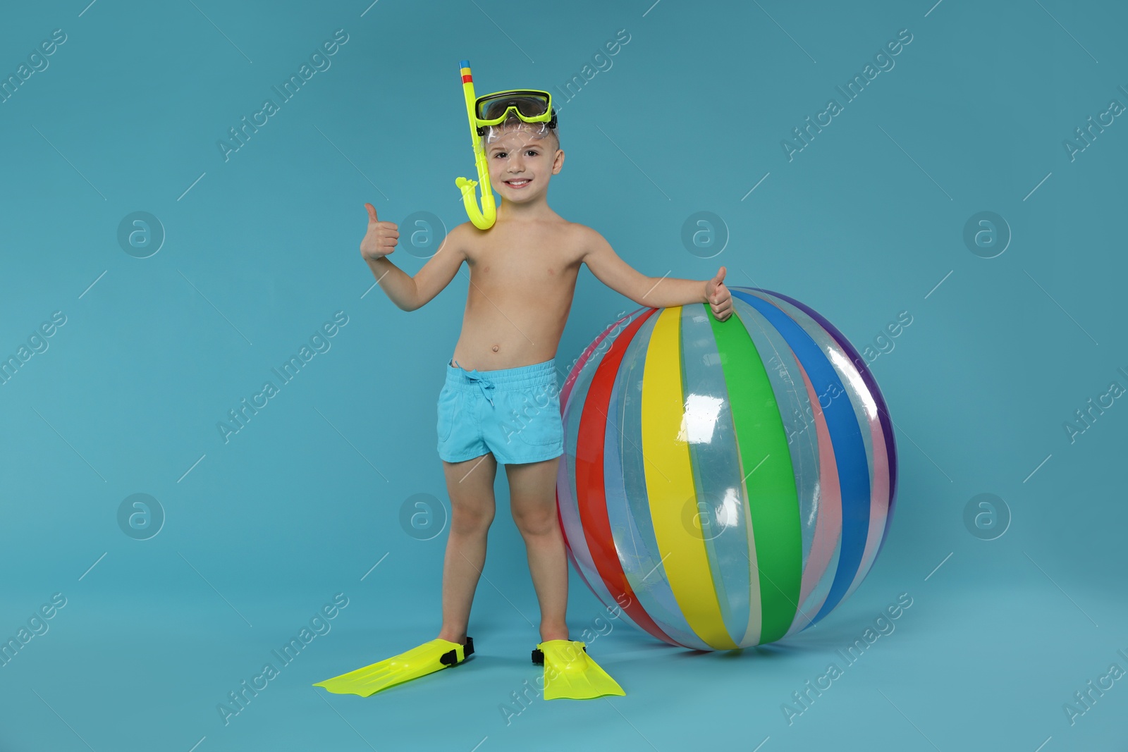 Photo of Cute little boy in beachwear with snorkeling equipment and inflatable ball showing thumbs up on light blue background