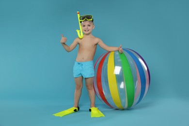 Photo of Cute little boy in beachwear with snorkeling equipment and inflatable ball showing thumbs up on light blue background