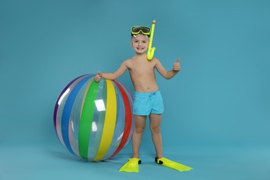 Photo of Cute little boy in beachwear with snorkeling equipment and inflatable ball showing thumbs up on light blue background