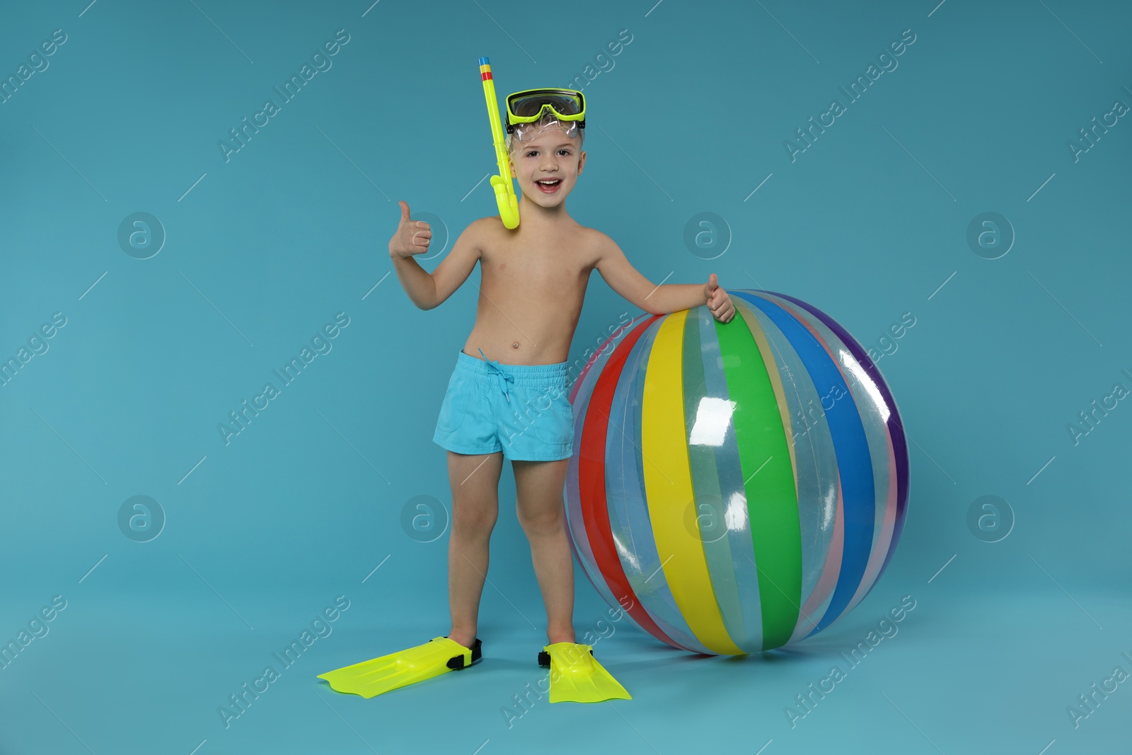 Photo of Cute little boy in beachwear with snorkeling equipment and inflatable ball showing thumbs up on light blue background