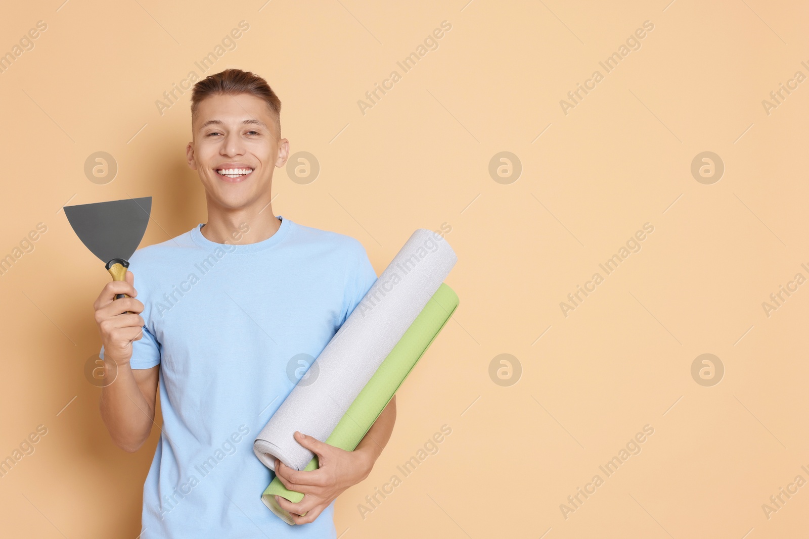 Photo of Young decorator with putty knife and rolls of wallpaper on beige background, space for text