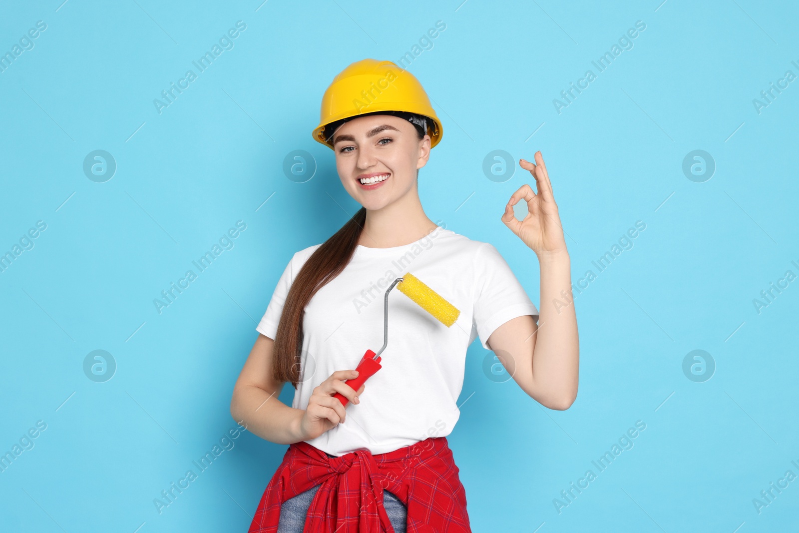 Photo of Portrait of young decorator with paint roller showing ok gesture on light blue background