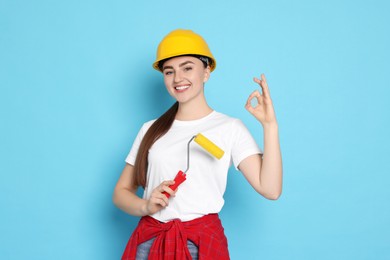 Photo of Portrait of young decorator with paint roller showing ok gesture on light blue background