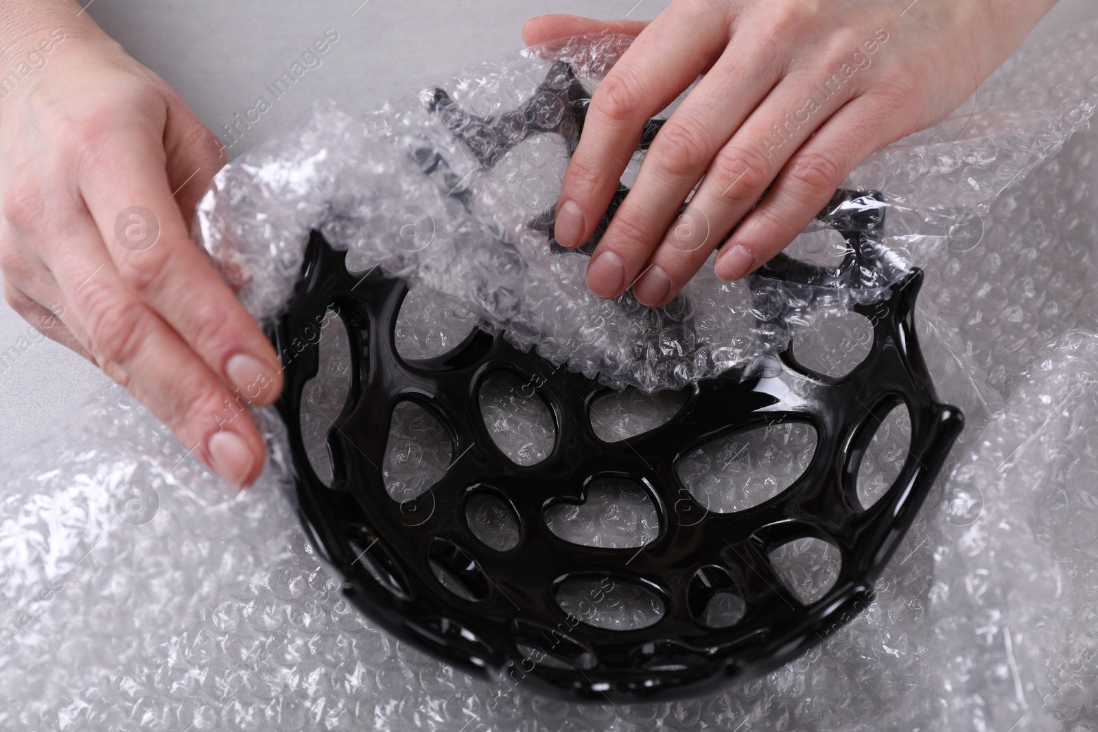 Photo of Woman covering ceramic bowl with bubble wrap at light grey table, closeup