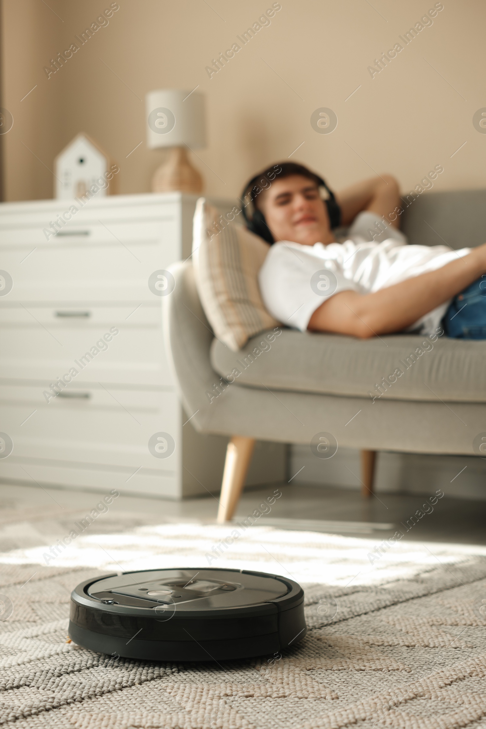 Photo of Robotic vacuum cleaner cleaning carpet while man relaxing on sofa, selective focus