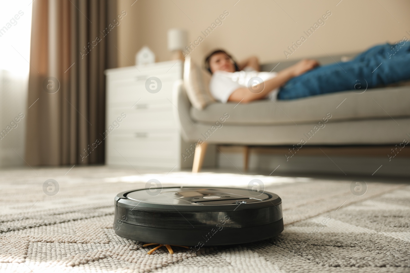 Photo of Robotic vacuum cleaner cleaning carpet while man relaxing on sofa, selective focus