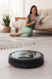 Photo of Young woman reading magazine at home, focus on robotic vacuum cleaner