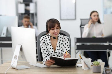 Photo of Saleswoman making notes while talking to client via headset at desk in office