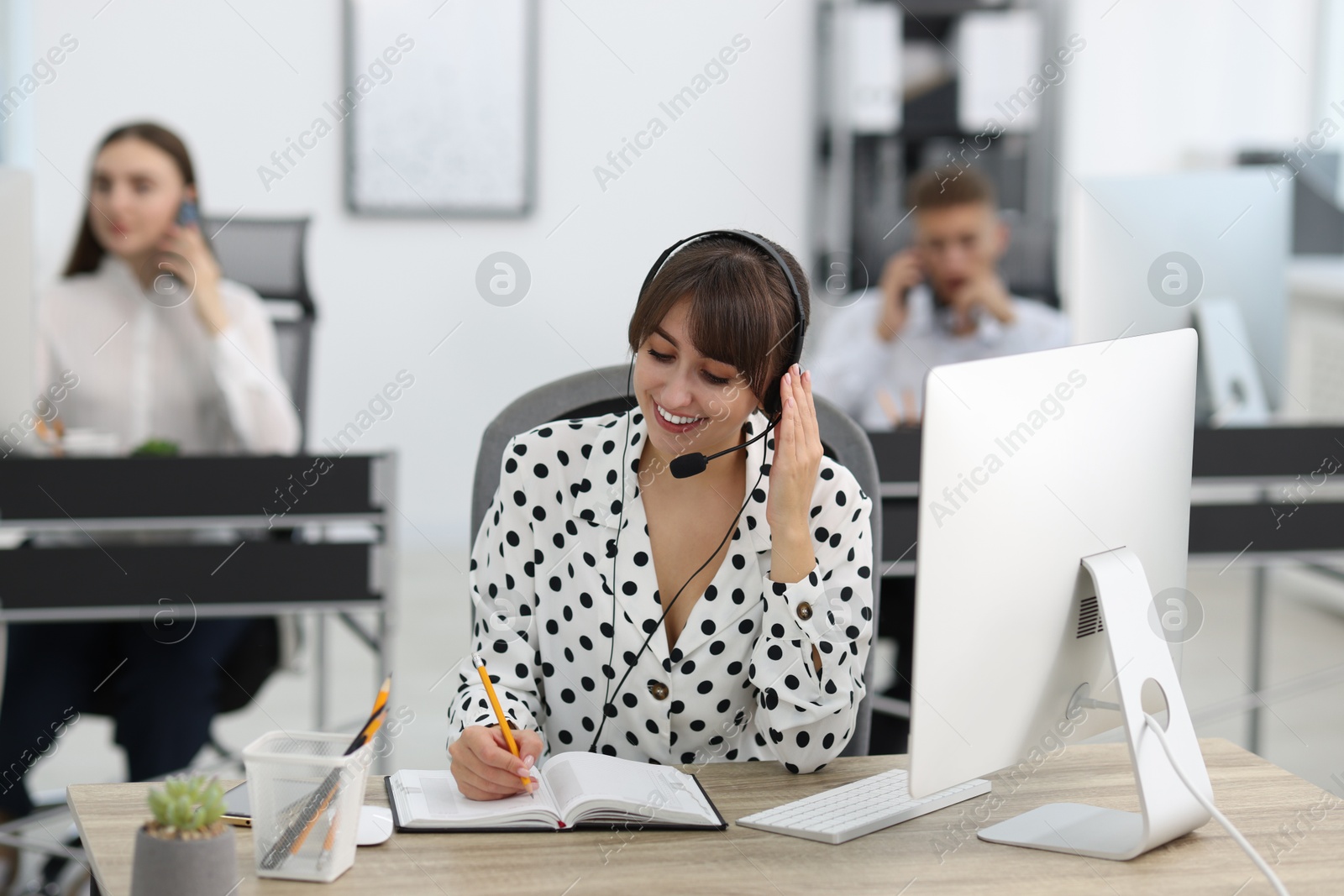 Photo of Saleswoman making notes while talking to client via headset at desk in office