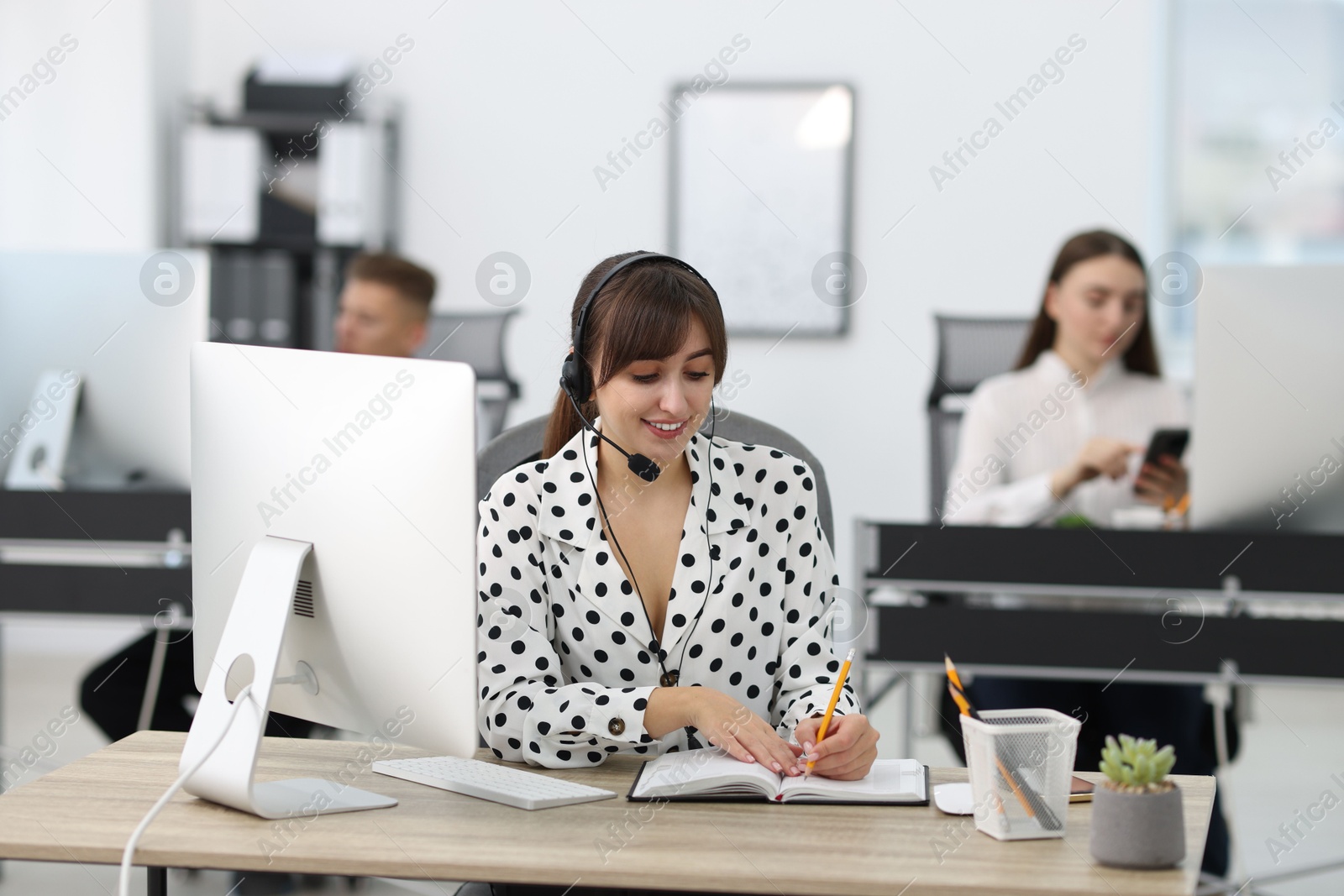 Photo of Saleswoman making notes while talking to client via headset at desk in office