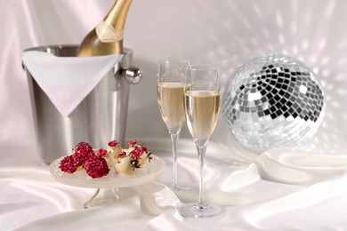 Photo of Champagne in glasses, ice bucket with bottle and tasty candies on white fabric