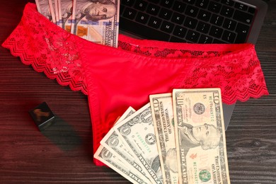 Photo of Sex work. Laptop with panties, dice and money on wooden surface, flat lay