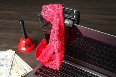 Photo of Sex work. Laptop with web camera, panties, bell and money on wooden surface, closeup