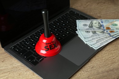 Photo of Sex work. Laptop with bell and money on wooden surface, closeup