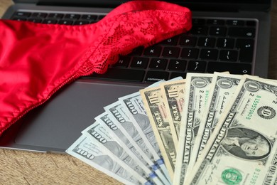 Photo of Sex work. Laptop with underwear and money on wooden surface, closeup