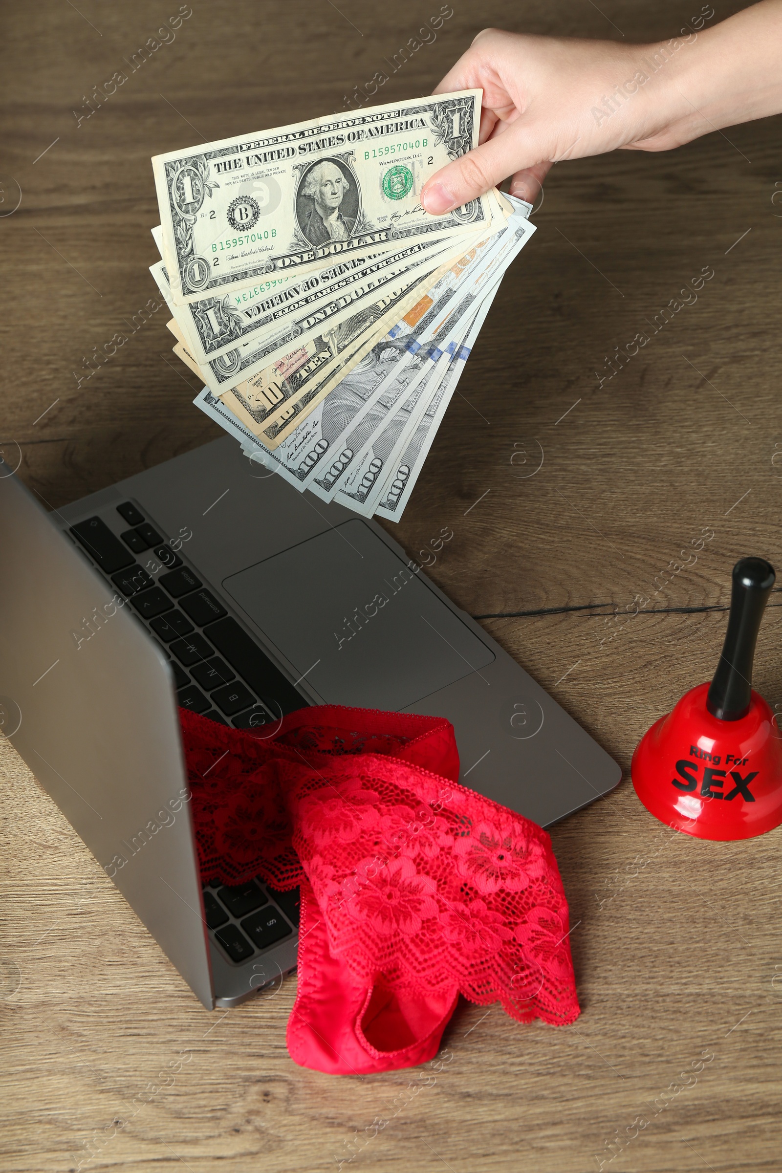 Photo of Sex work. Woman holding money in front of laptop with lingerie and bell on wooden surface, closeup