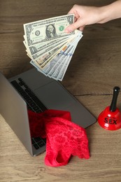 Photo of Sex work. Woman holding money in front of laptop with lingerie and bell on wooden surface, closeup