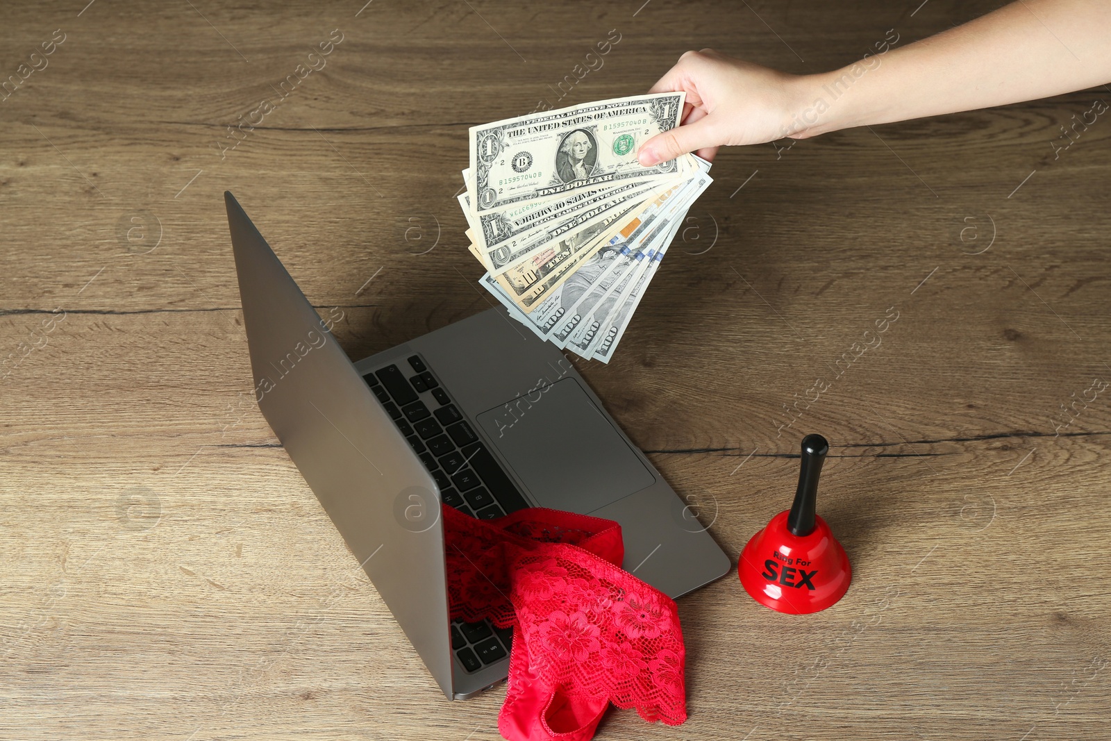 Photo of Sex work. Woman holding money in front of laptop with lingerie and bell on wooden surface, closeup