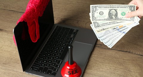 Photo of Sex work. Woman holding money in front of laptop with lingerie and bell on wooden surface, closeup