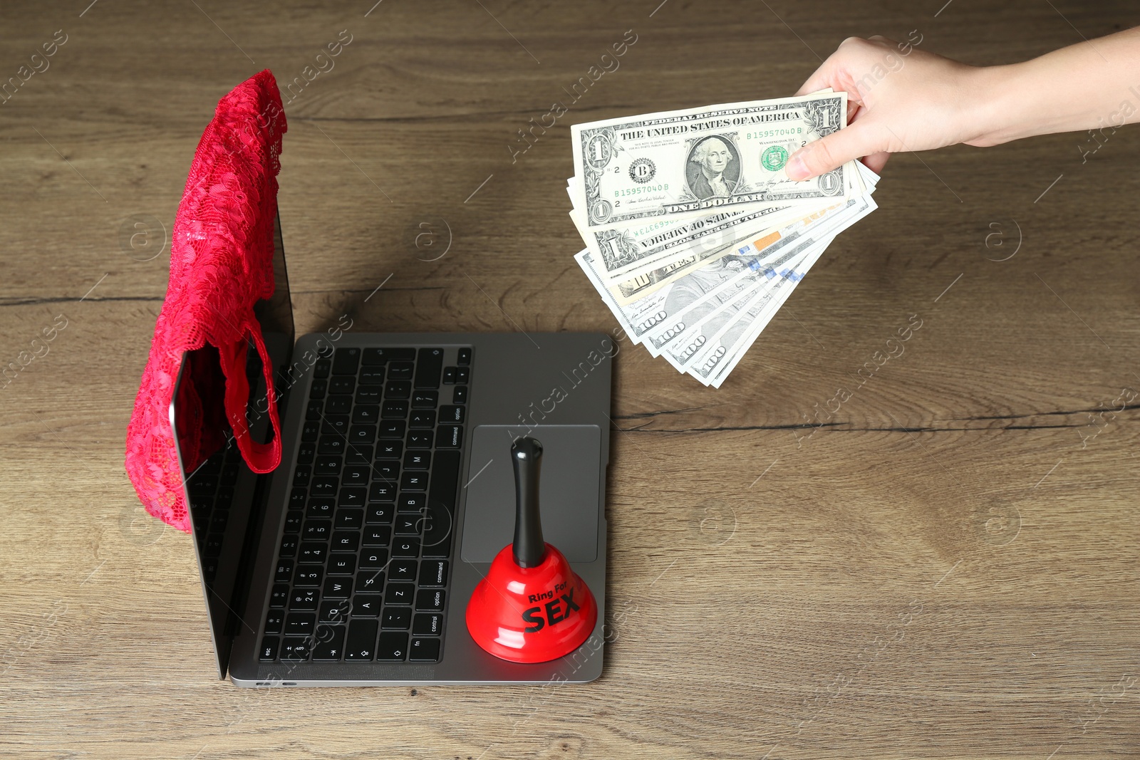 Photo of Sex work. Woman holding money in front of laptop with lingerie and bell on wooden surface, closeup