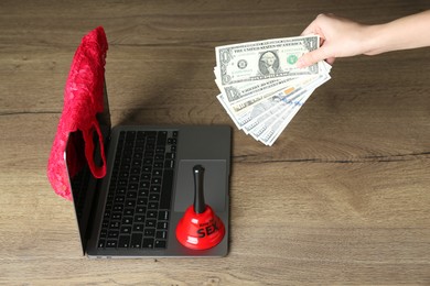 Photo of Sex work. Woman holding money in front of laptop with lingerie and bell on wooden surface, closeup