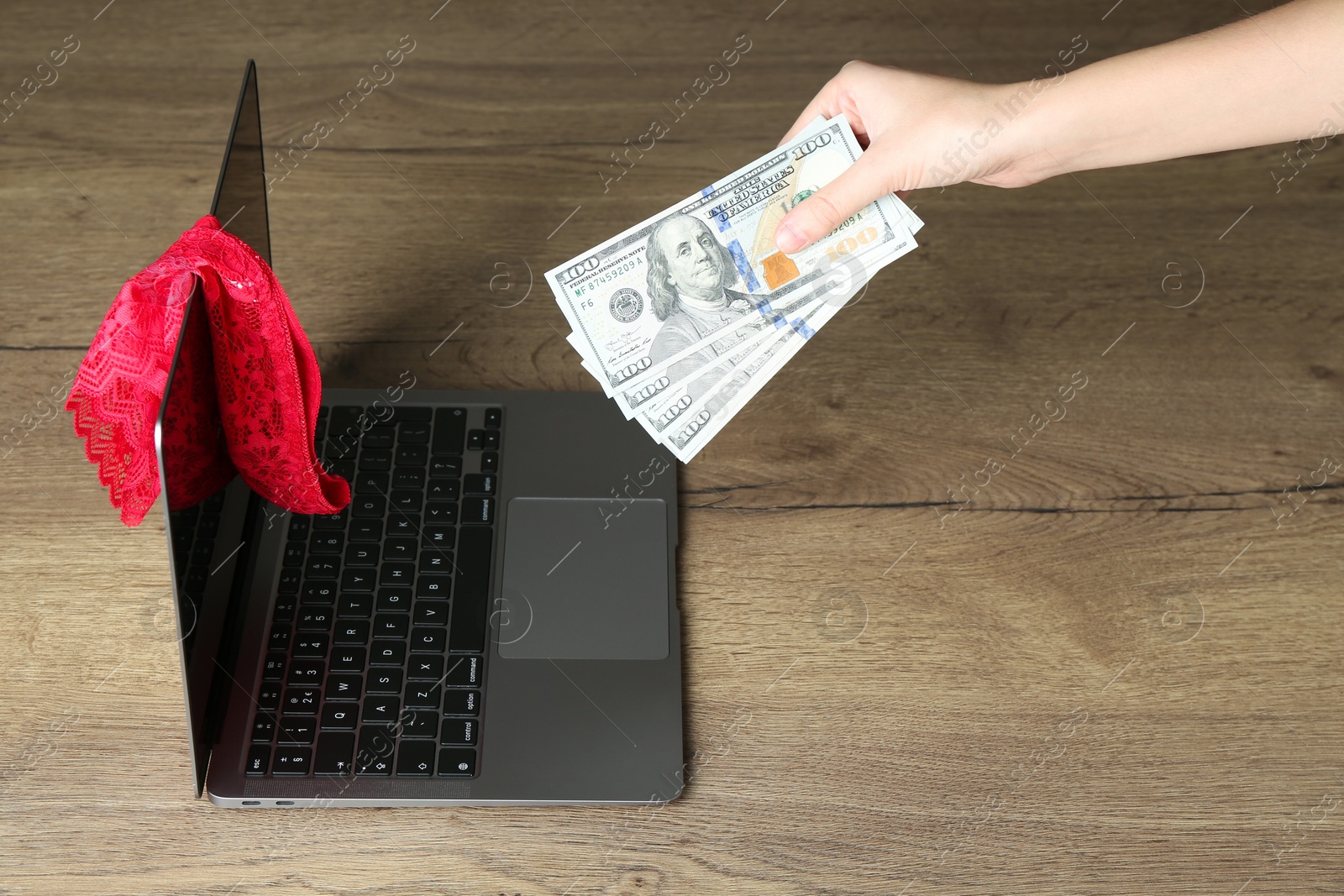 Photo of Sex work. Woman holding money in front of laptop with lingerie on wooden surface, closeup