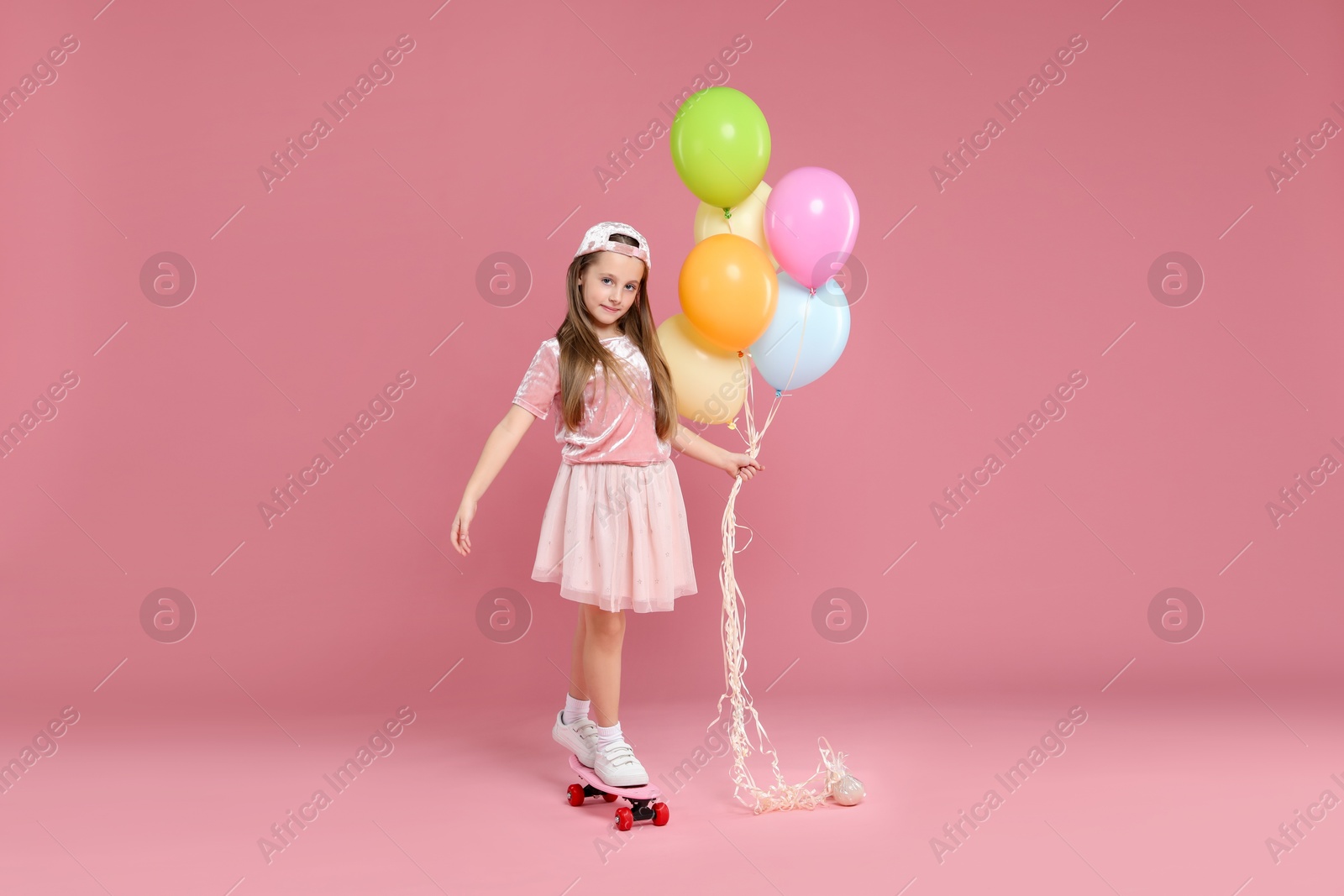 Photo of Little girl with colorful balloons standing on penny board against pink background