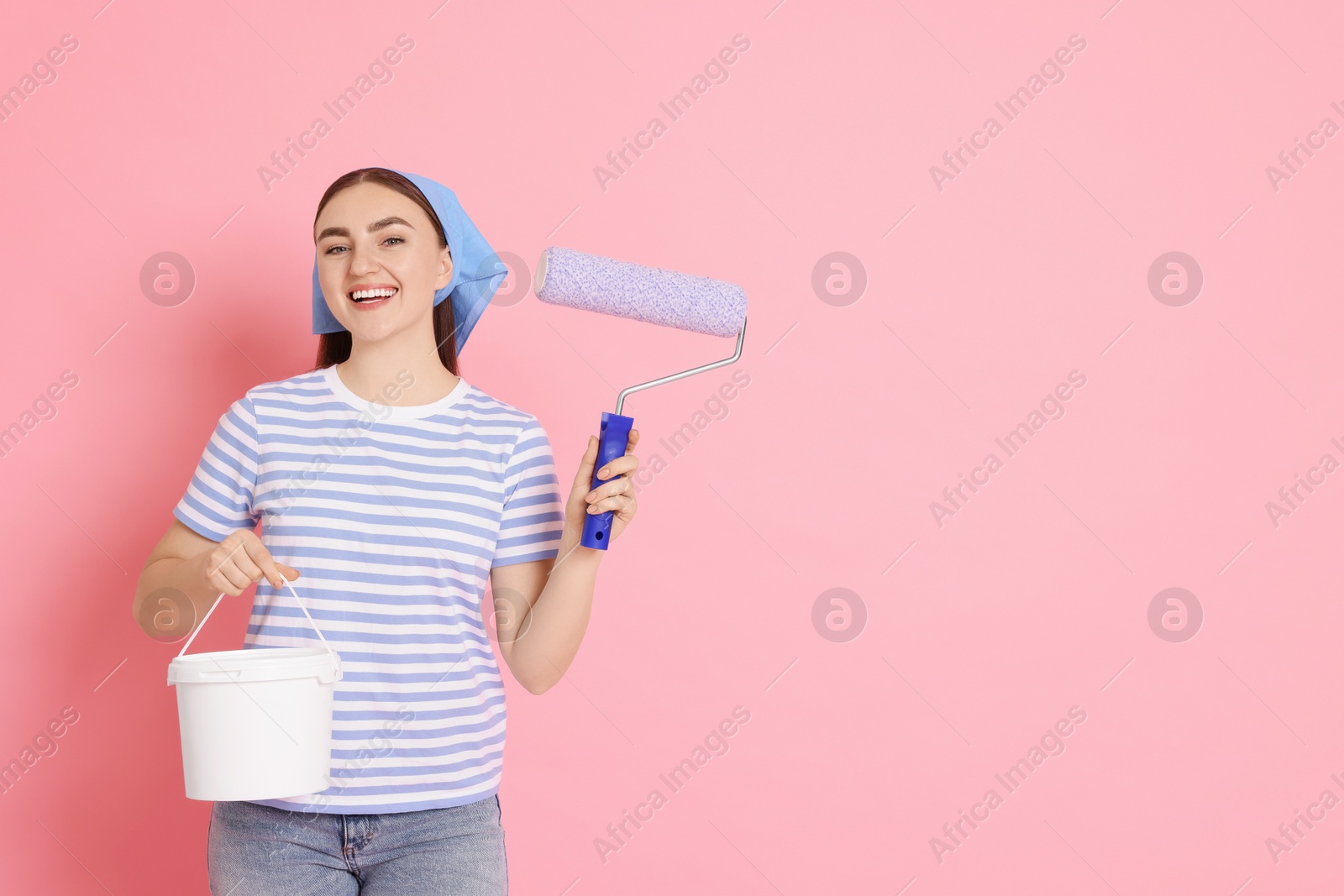 Photo of Portrait of young decorator with paint roller and bucket on pink background, space for text