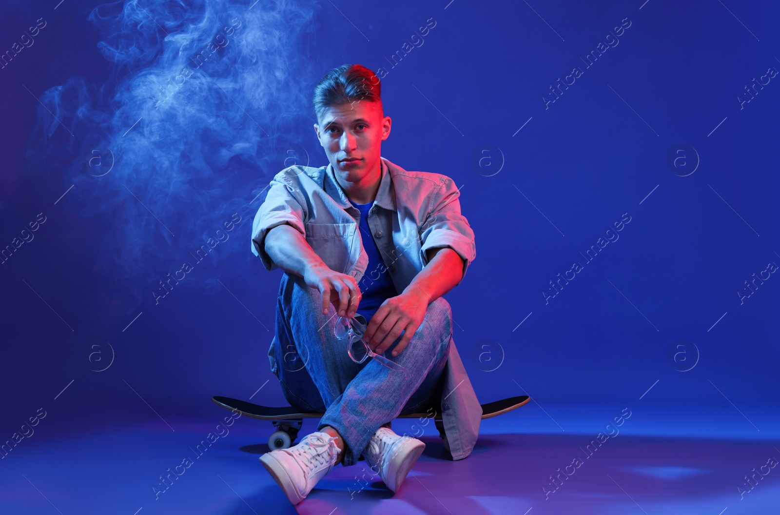 Photo of Handsome man sitting on skateboard in smoke and color lights against blue background