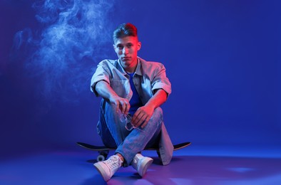 Photo of Handsome man sitting on skateboard in smoke and color lights against blue background