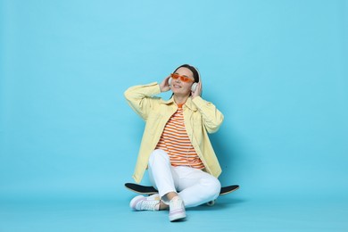 Smiling woman with headphones sitting on skateboard against light blue background