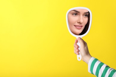 Beautiful woman looking in handheld mirror on yellow background, closeup. Space for text